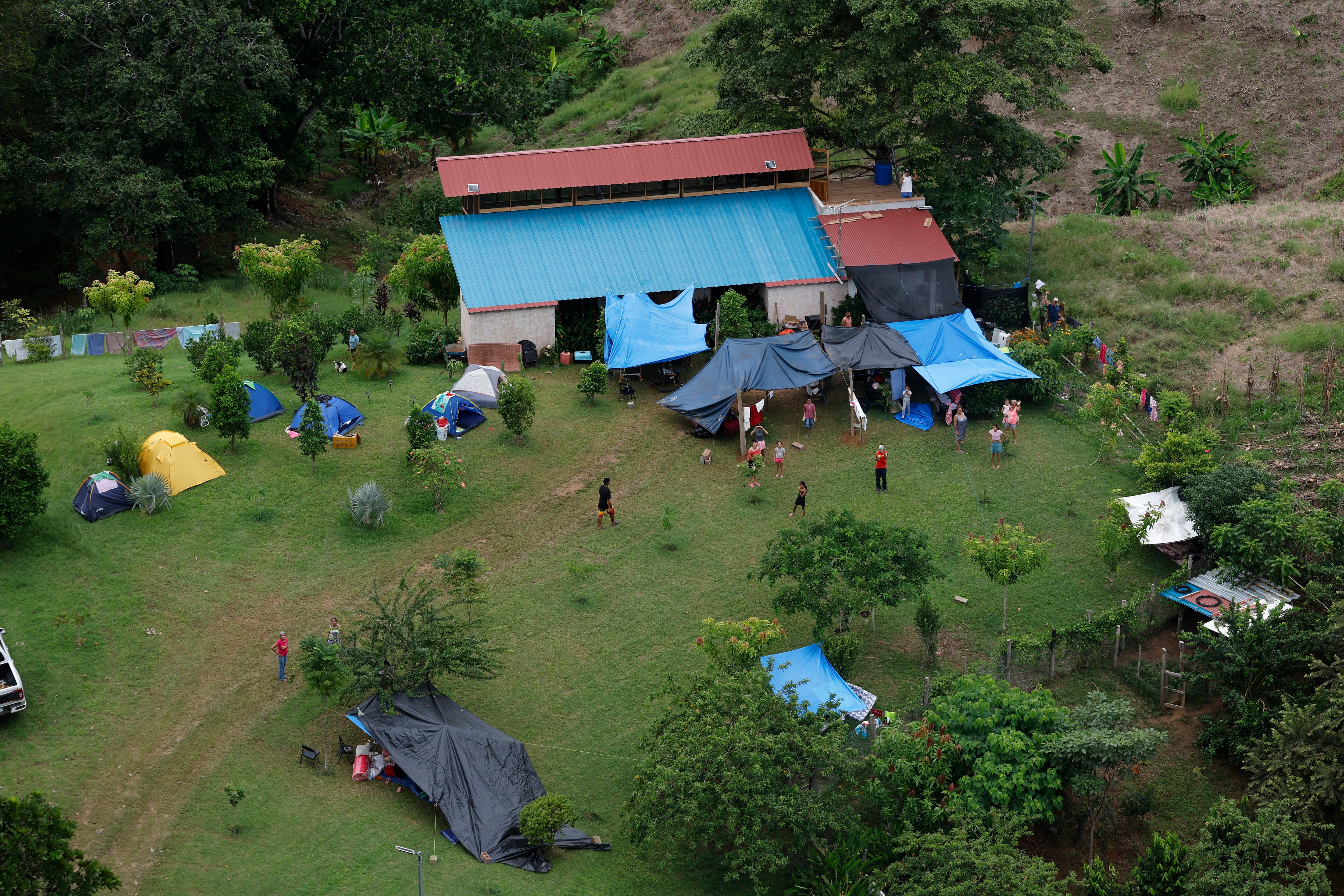 Varias personas permanecen en un campamento rural instalado sobre un terreno de hierba junto a una casa de techo azul.