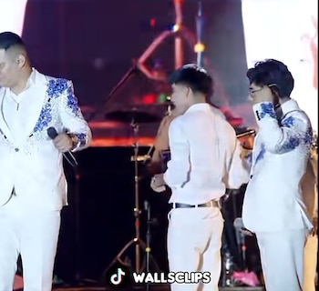 Tres músicos vestidos con trajes blancos con detalles de pedrería azul sostienen micrófonos sobre un escenario