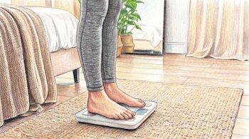 Primer plano de pies descalzos sobre una báscula blanca digital en un tapete beige, con parte de una pierna vestida de gris en una habitación con muebles.