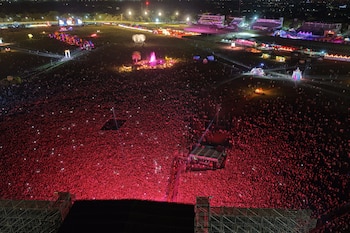 Vista aérea nocturna de un festival al aire libre con una enorme multitud bajo luces rojas. Se ven escenarios, torres de iluminación y otras estructuras coloridas