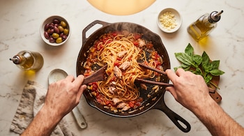 Vista cenital de manos mezclando pasta con tomate y atún en una sartén, junto a recipientes con aceitunas, ajo, aceite y albahaca.