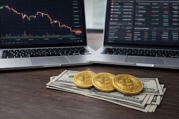 Primer plano de tres monedas Bitcoin doradas y unos billetes de dólar acostados sobre una superficie de madera, con dos laptops abiertas de fondo.
