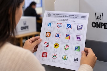 Manos de una persona sostienen una cédula de papel con varios logotipos de partidos políticos frente a un módulo electoral