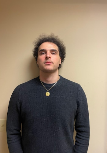 Hombre joven con cabello rizado oscuro, suéter de punto oscuro y collar dorado con un medallón. Fondo de pared beige claro con un interruptor