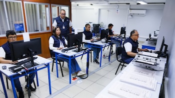 Trabajadores con chalecos azules operan computadoras en una oficina mientras supervisores observan y hay pilas de documentos en las mesas
