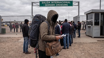 Persona encapuchada con mochila y una bolsa, sosteniendo papeles, de espaldas, ante una fila de gente y un cartel verde de "Salida Internacional".