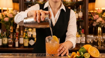 Primer plano de un bartender, con camisa blanca y chaleco oscuro, vertiendo un cóctel anaranjado desde una coctelera a un vaso con hielo y limón, rodeado de cítricos frescos.