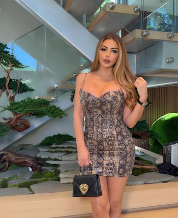 Larsa Pippen posa con un vestido corto con estampado de piel de serpiente y un bolso negro, frente a una escalera moderna de madera y vidrio con plantas