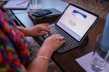 Manos escribiendo en un teclado de netbook. La pantalla muestra un documento sobre la Política Nacional de Desarrollo de las Mujeres. Un teléfono y un vaso en la mesa