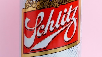 Primer plano del logo 'Schlitz' en cursiva blanca con borde negro y dorado sobre un fondo rojo vibrante de una lata, con detalles de lúpulo y fondo rosa