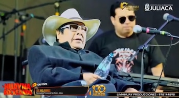 Un hombre con sombrero claro, gafas y abrigo sostiene una botella en un escenario musical frente a un micrófono y junto a un tecladista