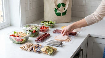 Mesa de cocina blanca con varios envases de alimentos listos para consumir: ensaladas, cárnicos, snacks. Una mano toma un producto. Al fondo, una bolsa de tela con símbolo de reciclaje.