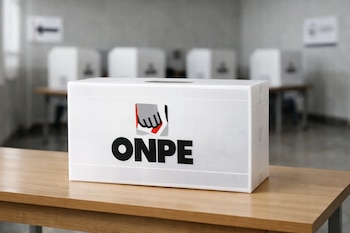Urna electoral blanca con logo ONPE en una mesa de madera. Al fondo desenfocado, cabinas de votación y señalética en un centro electoral.