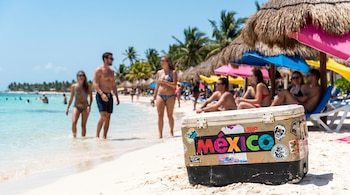 Hielera de color beige y negro con calcomanías, incluyendo el logo de México, en la arena de una playa con mar turquesa, palmeras y personas.