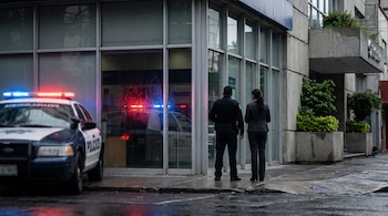 Un policía y una mujer están de espaldas frente a un gran ventanal de un banco, junto a una patrulla con luces azules y rojas encendidas.