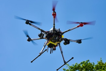 Dron hexacóptero negro con seis rotores en movimiento, cuatro negros y dos rojos, y tren de aterrizaje metálico, suspendido en un cielo azul brillante