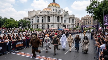 Personas disfrazadas de personajes de terror corren por una calle vallada frente al Palacio de Bellas Artes ante una multitud.