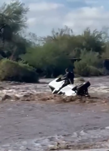 Una camioneta blanca volcada en la corriente de un río caudaloso con una persona sobre el vehículo y vegetación al fondo