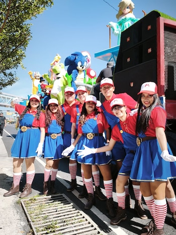 Siete personas con disfraces de overol azul, camiseta roja y gorra roja con la letra M. Detrás, una carroza con figuras de Sonic, Yoshi y Rosalina. Día soleado