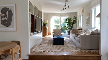 Un salón iluminado con sofá beige, mesa negra, alfombra estampada, estantería integrada y una planta verde junto a la ventana.