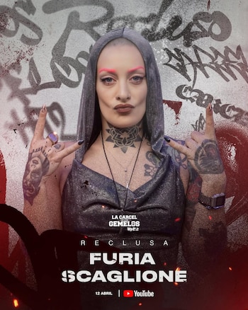 Mujer con cabeza rapada, cejas rosadas y tatuajes en cuello y brazos, viste una sudadera con capucha gris y hace gestos de rock and roll ante un grafiti