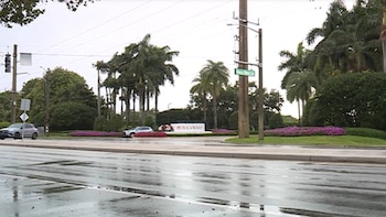 Vista de una intersección con asfalto mojado, dos coches, un semáforo, postes eléctricos, palmeras y una señal de 'BOCAWEST' bajo un cielo nublado