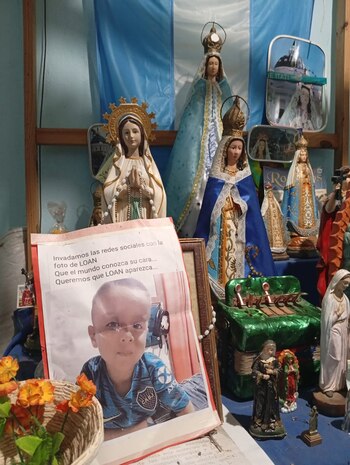 Altar doméstico con varias figuras de la Virgen María y santos, un cartel con la foto del niño Loan Peña en primer plano, una bandera celeste y blanca de fondo y flores artificiales