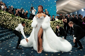 Megan Thee Stallion eligió un vestido plateado ajustado al cuerpo, completamente bordado con pedrería y lentejuelas, con escote profundo en forma de corazón y tajo lateral alto que deja una pierna al descubierto. La prenda está acompañada por un abrigo de gran volumen en piel sintética blanca, que cae hasta el suelo y genera una silueta majestuosa por su longitud y textura