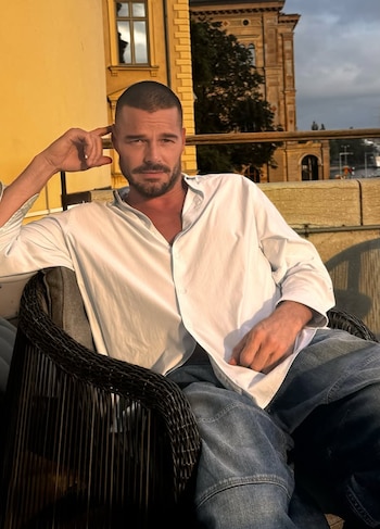Hombre con barba y cabello corto, camisa blanca, jeans azules, sentado en silla de mimbre oscura en exterior con edificios amarillos de fondo
