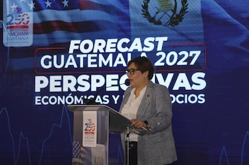 Mujer en podio con micrófono. Pantalla de fondo azul con texto "Forecast Guatemala 2027 Perspectivas Económicas y Negocios", banderas de Estados Unidos y Guatemala, y logo de AmCham