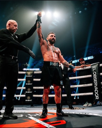 Un hombre con barba y tatuajes levanta el brazo en un ring de combate. Un árbitro a su lado. Se observan cuerdas, público y logos de marcas