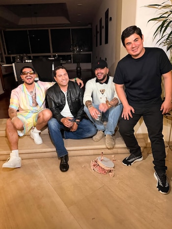 Cuatro hombres sonríen y posan sentados en un suelo de madera clara. Visten ropa casual. Un pastel parcialmente destrozado está en el suelo frente a ellos