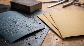 Una lija de taco, lijas de agua grises con gotas, varias lijas secas marrones y dos herramientas de metal sobre una mesa de madera.