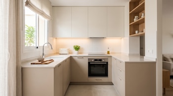 Cocina en forma de U con muebles arena, mesada blanca, fregadero bajo una ventana, placa de cocción, horno y estantería de madera en la pared.