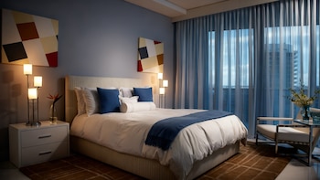 Dormitorio moderno con cama, colchón y sábanas blancas. Almohadas azules, dos mesitas de noche con lámparas encendidas y arte abstracto en paredes azules