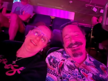 Dos personas, un hombre con bigote y una mujer con gafas, se toman una selfie en un recinto con iluminación púrpura