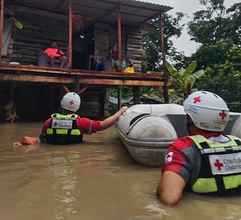 Tres rescatistas con uniformes de la Cruz Roja, cascos y chalecos, junto a un vehículo de rescate blanco. El fondo muestra un paisaje montañoso y húmedo con vegetación