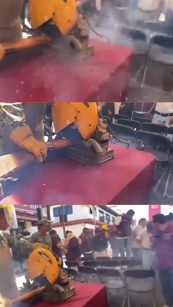 Militar usando una sierra de corte amarilla para desarmar un arma de fuego sobre una mesa roja. Hay chispas, humo, personas y sillas en el fondo