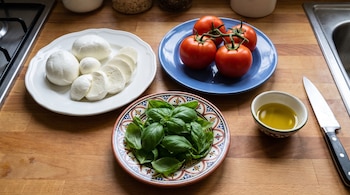 Tres platos sobre una tabla de madera con queso mozzarella, tomates rojos y hojas de albahaca verde, junto a un recipiente con aceite de oliva y un cuchillo.