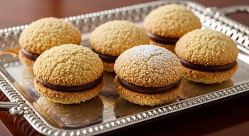 Seis galletas redondas de avellana con relleno de chocolate en una bandeja de plata, una de ellas espolvoreada con azúcar glas.