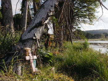 Dos cruces, una de madera decorada con espejos y otra rosa con un rosario y flores blancas, al pie de un árbol grande. Hay vegetación frondosa y un río