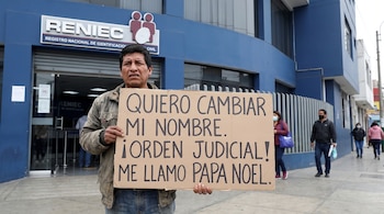 Un hombre de tez morena sostiene un cartel de cartón frente a un edificio azul con el logotipo de RENIEC, mientras personas transitan en la calle.