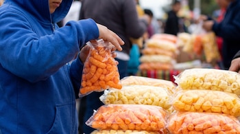 Niño con sudadera azul sostiene una bolsa de snacks naranjas. Hay pilas de bolsas con snacks naranjas y amarillos en un puesto. Fondo con personas.