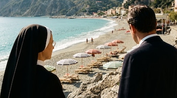 Monja y hombre de traje de espaldas mirando una playa de arena con mar, sombrillas a rayas, reposeras, pocas personas y edificios costeros.