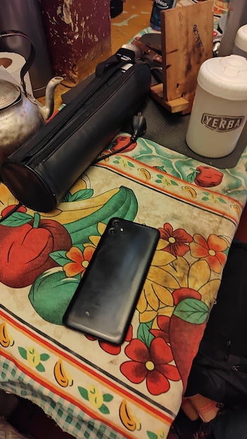 Un teléfono celular negro sobre un mantel floreado, junto a una pava metálica, un recipiente de yerba y un estuche negro