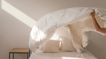 Una mano está ajustando un edredón blanco sobre una cama con sábanas y almohadas blancas en un dormitorio, con un rayo de sol diagonal en la pared.