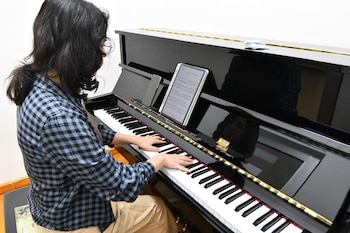 Una persona con cabello oscuro y camisa a cuadros azul y negro toca un piano de cola negro con partituras digitales en una tableta sobre el atril