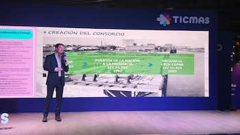Hombre vestido de traje negro y camisa blanca hablando en un micrófono sobre un escenario frente a una gran pantalla azul con gráficos de un puerto antiguo y texto