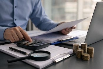 Manos de una persona usando una calculadora sobre papeles con gráficos, una lupa, pilas de monedas y una computadora portátil en un escritorio.