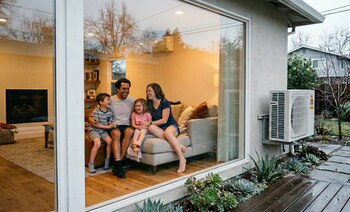 Vista a través de una ventana de una familia sonriente sentada en un sofá dentro de una casa, con una bomba de calor blanca en la pared exterior
